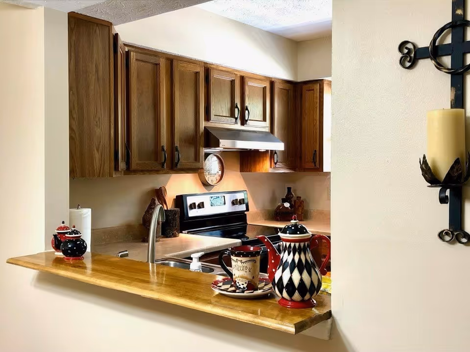 Interior view of a kitchen with wooden cabinets, a stove with a range hood, a sink, and a wooden countertop bar. On the countertop are decorative items including a teapot, a cup and saucer, and a small container. A wall-mounted candle holder with a yellow candle is visible on the right side.