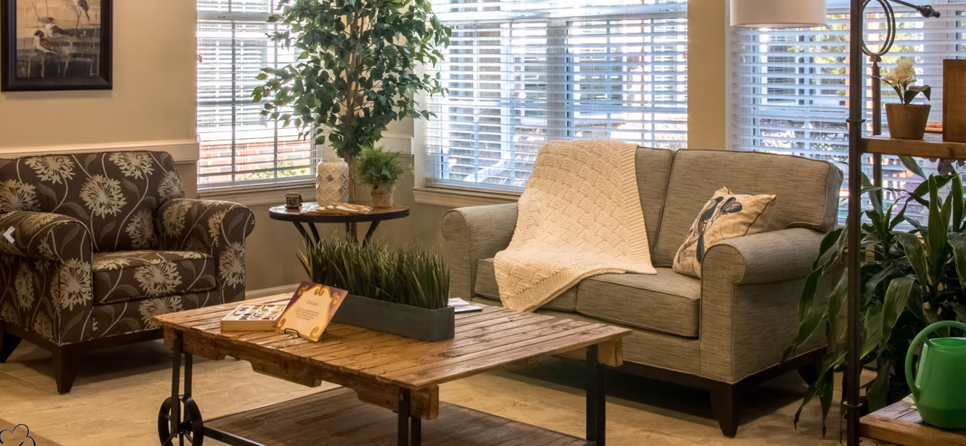 A cozy living room area with a gray upholstered loveseat draped with a cream knitted blanket and a decorative pillow, a floral patterned armchair, a wooden coffee table with a rectangular planter of green grass, and a round side table with potted plants. Large windows with white blinds allow natural light to fill the space.