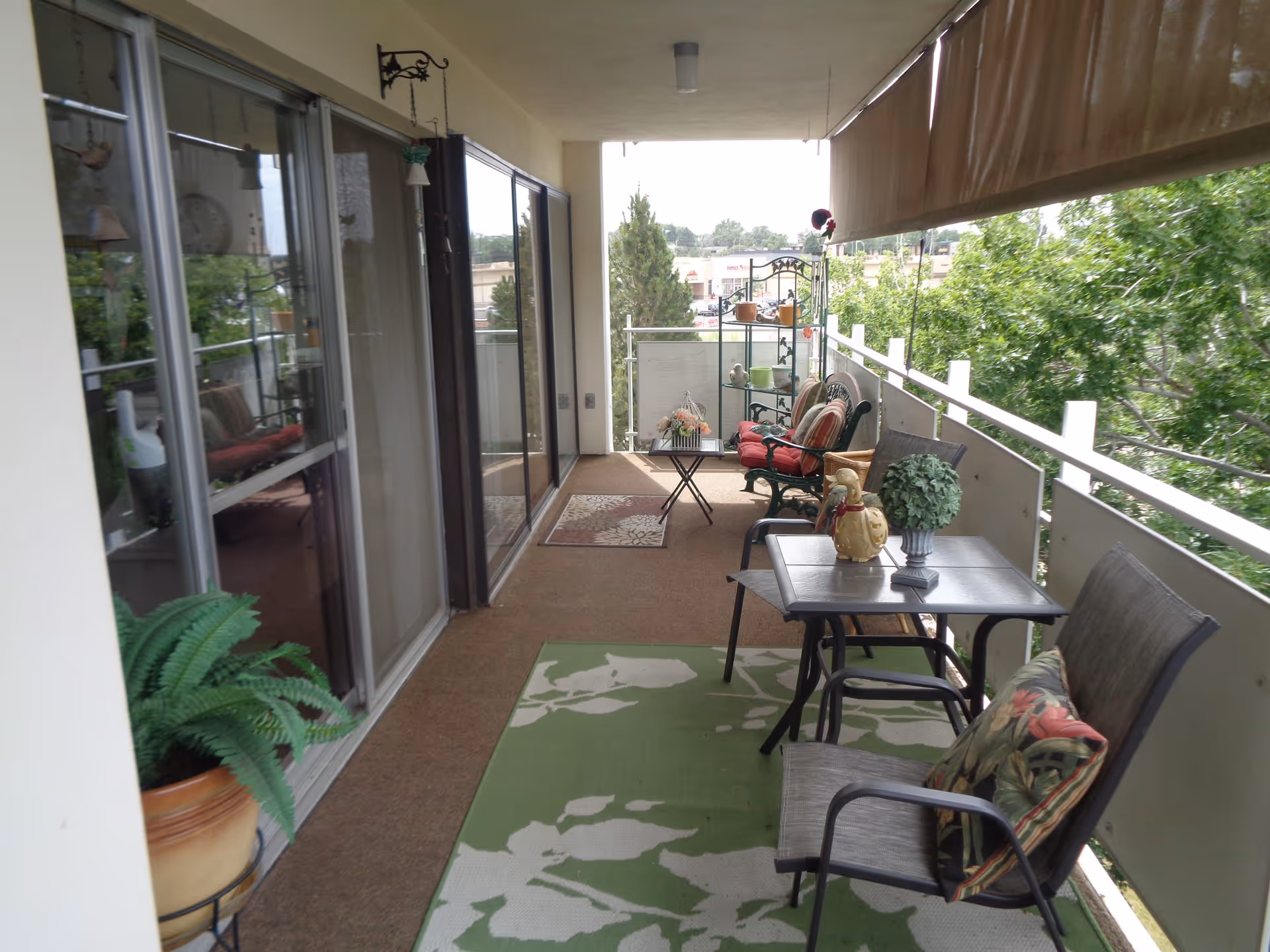 A covered balcony with outdoor seating including chairs with cushions, a small table with decorative items, potted plants, and a green patterned rug. The balcony overlooks trees and a distant view of buildings.