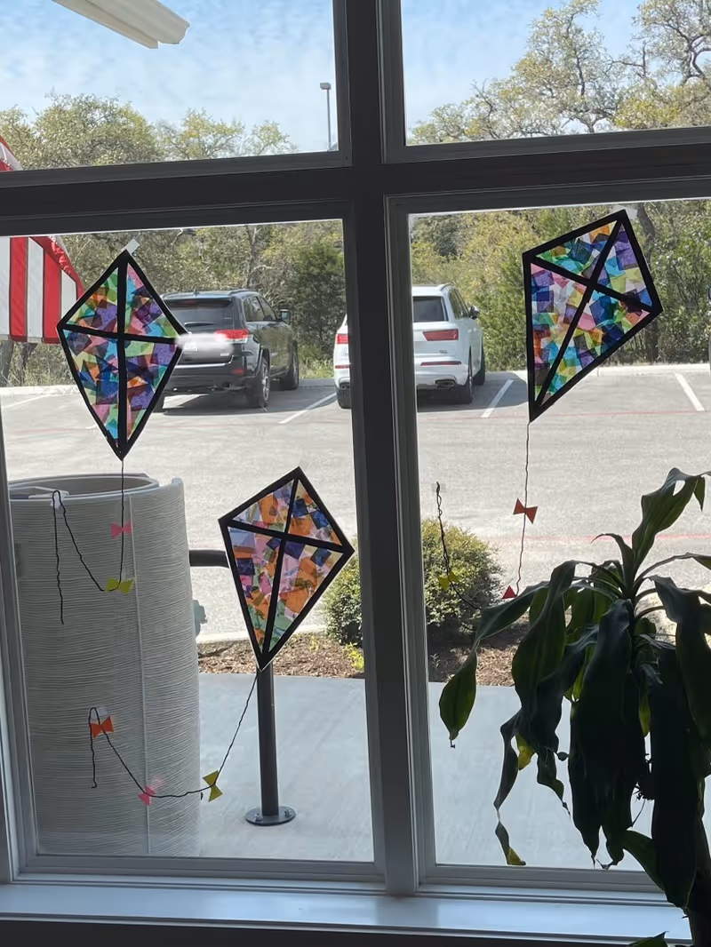 View through a window showing three colorful paper kites taped to the glass. Outside, there is a parking lot with two parked cars and some greenery. Inside, a large green plant is visible near the window.