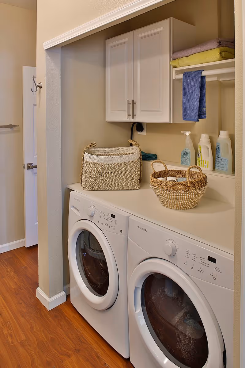 Laundry area with a front-loading washing machine and dryer side by side, a countertop above them holding two woven baskets, and a wall-mounted cabinet with folded towels and cleaning supplies on a shelf underneath.