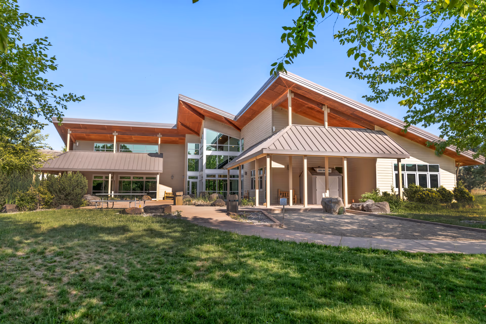 Exterior view of a modern senior living facility building with large windows, a sloped roof, surrounded by green grass and trees under a clear blue sky.