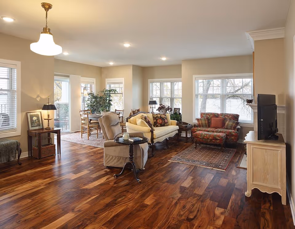 A spacious living room with polished wooden floors, featuring a beige armchair with a small round side table, a yellow upholstered sofa with floral cushions, and a floral-patterned loveseat with a matching ottoman. The room has large windows letting in natural light, a TV on a light-colored wooden stand, and a dining area with wooden chairs and a table in the background. There are also several lamps and a potted plant near the dining area.