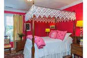 Bright bedroom with a white four-poster canopy bed against pink and red walls, wooden nightstands, lamps, and a window with yellow curtains.