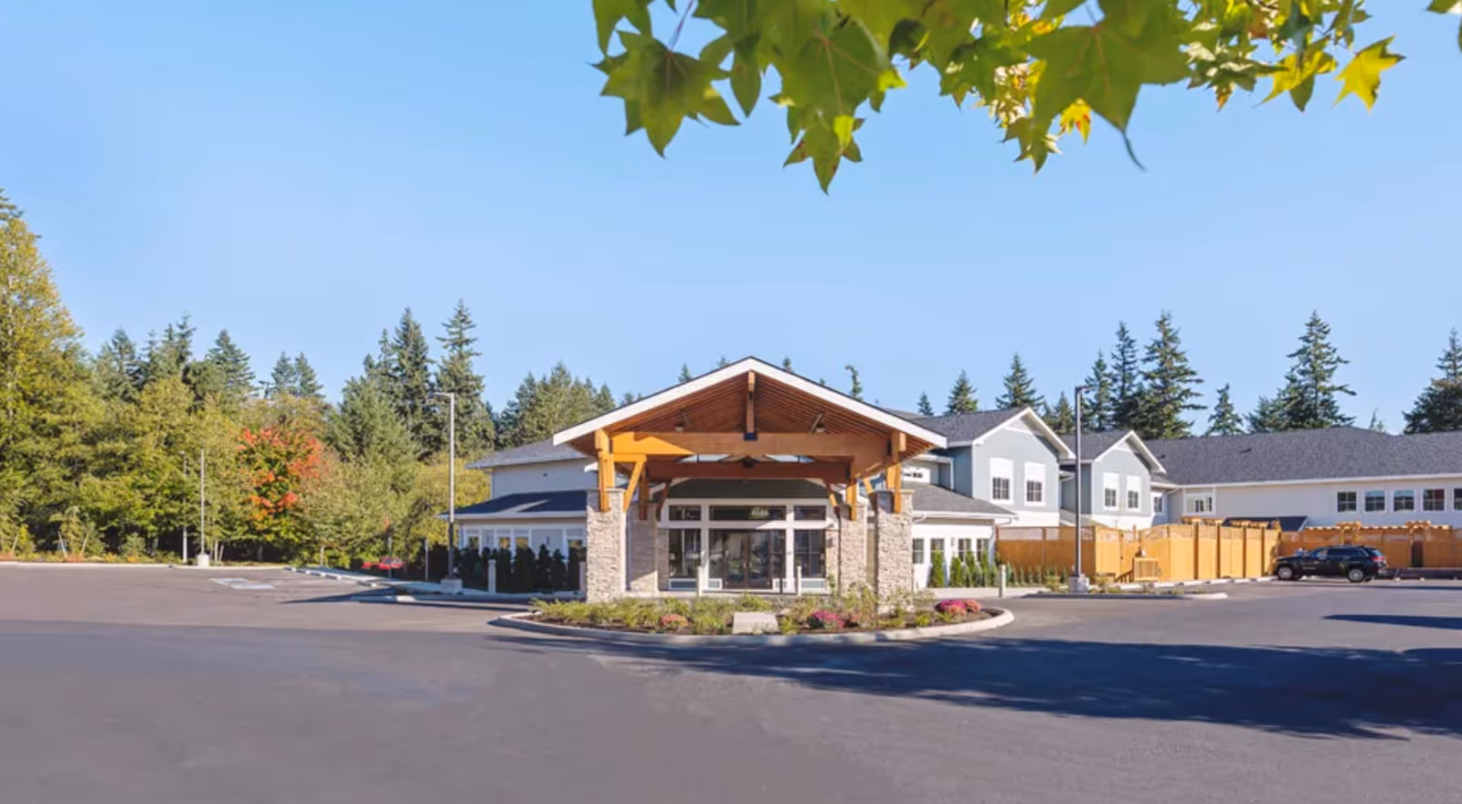 Exterior view of Mukilteo Memory Care facility showing a large building with a covered entrance, surrounded by a parking lot and trees under a clear blue sky.