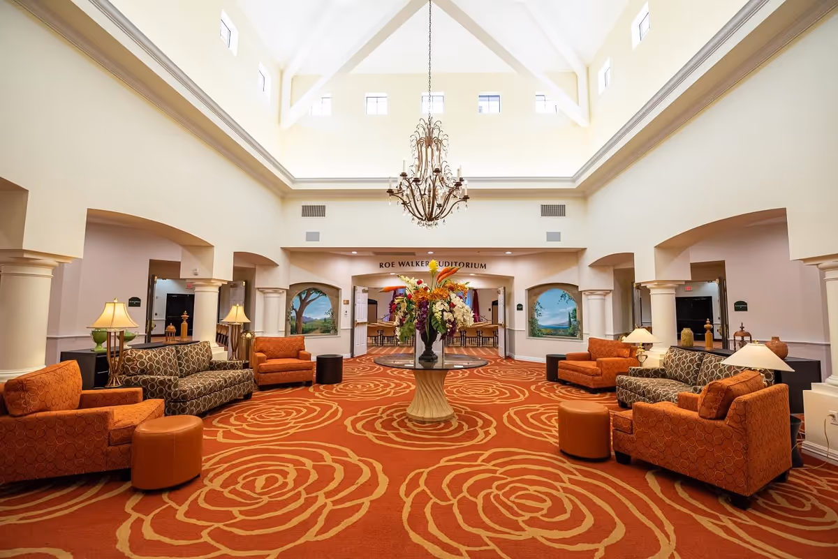 Large bright lobby/lounge with a chandelier, patterned red carpet, multiple seating areas and a central floral arrangement.