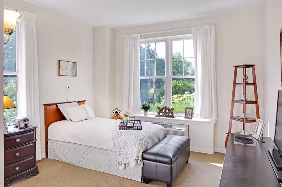 A bright and cozy bedroom with a single bed featuring white bedding and a light gray bed skirt. The room has beige carpet, a wooden nightstand with a lamp and clock, a large window with white curtains letting in natural light, and a black leather bench at the foot of the bed. There is a wooden corner shelf with decorative items and a flat-screen TV on a dark wooden stand.