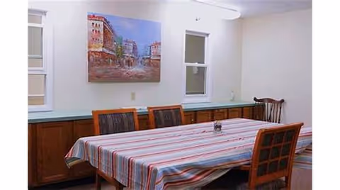 A dining room with a rectangular table covered by a striped tablecloth. Four wooden chairs with cushioned seats surround the table. The room has two windows with blinds, a wooden sideboard along the wall, and a painting of a cityscape hanging above the sideboard.