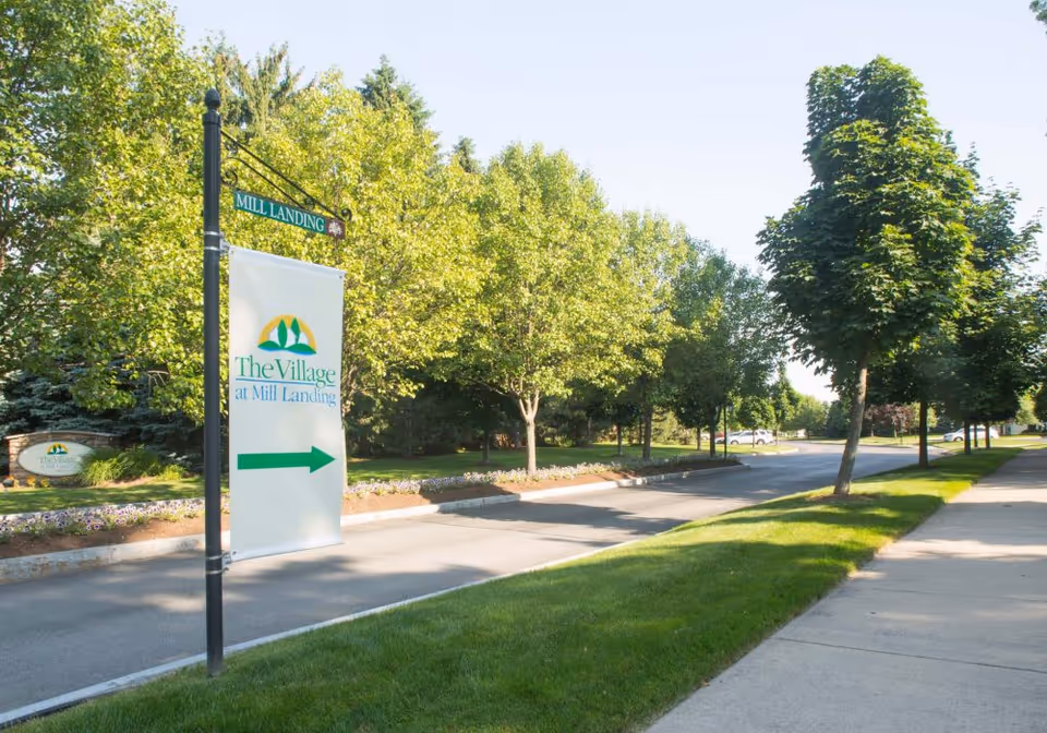 A sunny street view with a sidewalk and green grass on the right side, lined with trees. A black street lamp post on the left displays a hanging sign with the text 'The Village at Mill Landing' and a green arrow pointing right. In the background, more trees and parked cars are visible.