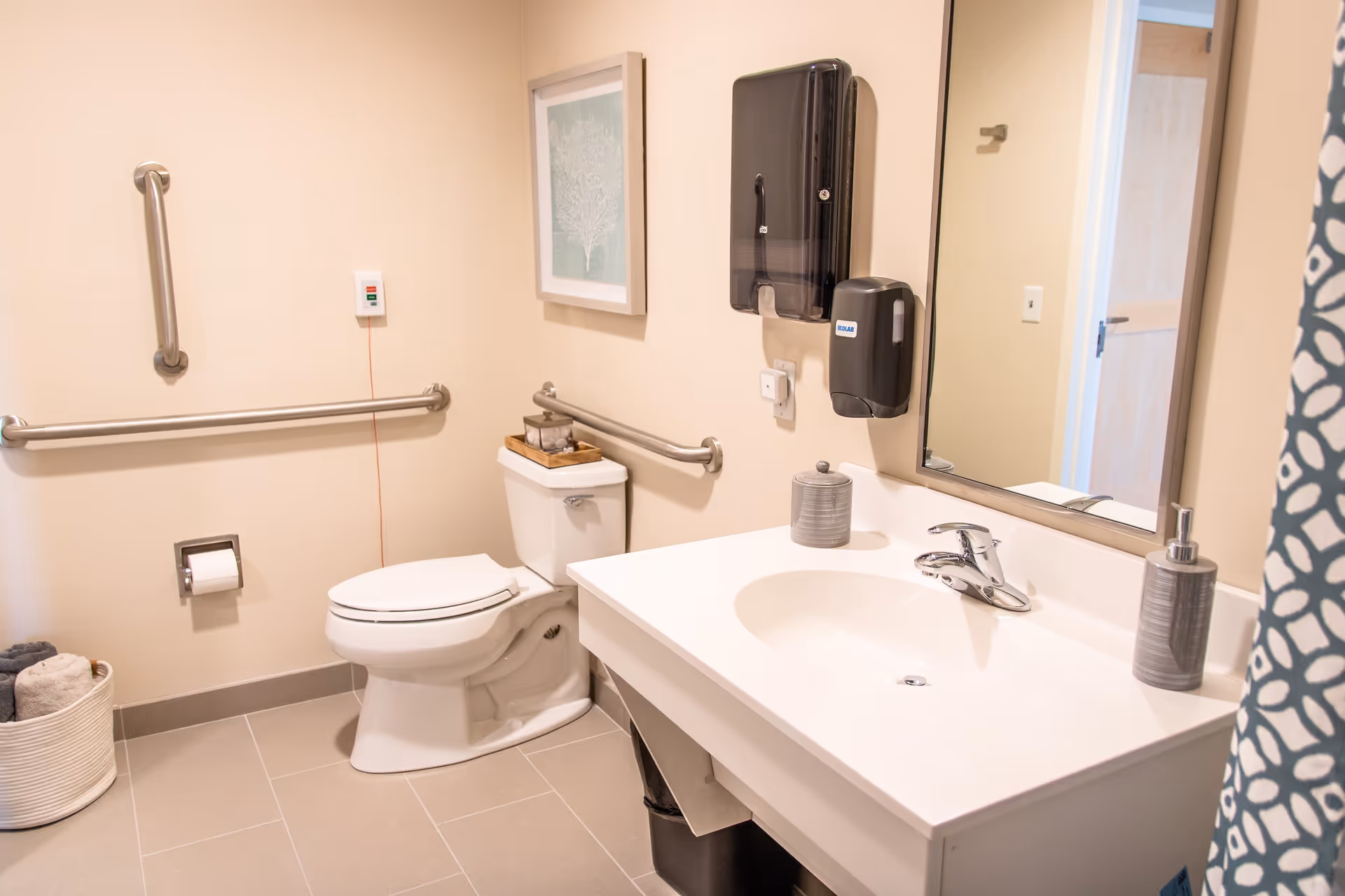 A clean and accessible bathroom with beige walls and light gray tiled floor. The bathroom features a white toilet with grab bars on the walls for support, a white sink with a modern faucet, a large mirror above the sink, a soap dispenser, and a paper towel dispenser mounted on the wall. There is a framed artwork on the wall above the toilet and a basket with rolled towels on the floor.