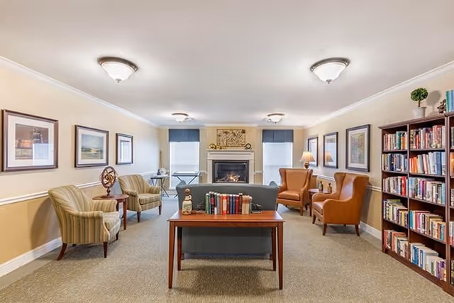 A cozy living room in a senior living facility featuring a fireplace centered on the far wall between two windows with blue valances. The room has a mix of seating including two striped armchairs on the left, two brown leather armchairs on the right, and a blue sofa in the middle facing the fireplace. A wooden table behind the sofa holds a collection of books and decorative items. The walls are adorned with framed artwork, and a tall bookshelf filled with books stands on the right side of the room. The ceiling has multiple light fixtures providing warm lighting.