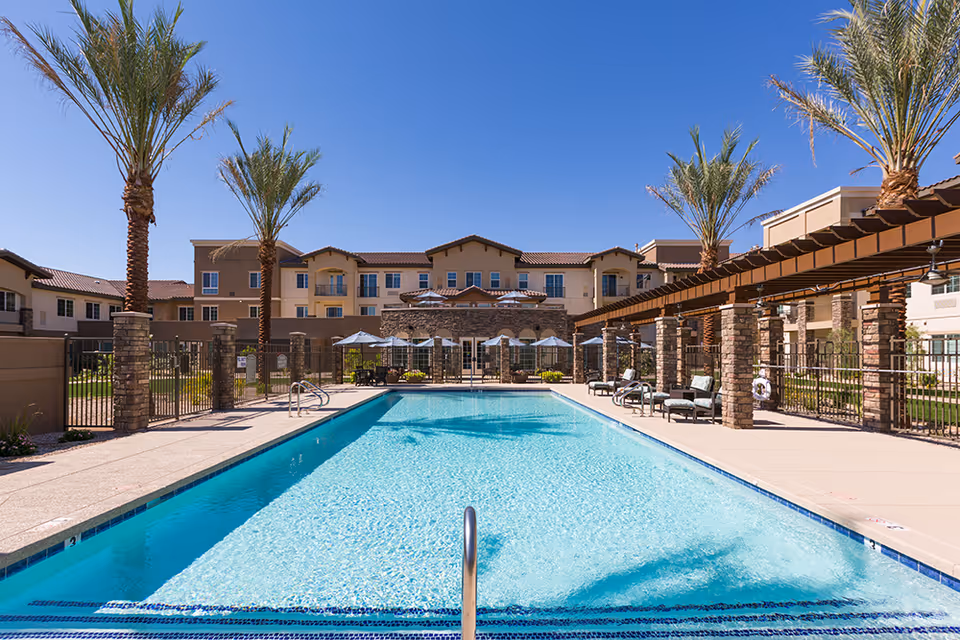 Outdoor swimming pool area at The Enclave at Chandler Senior Living with clear blue water, surrounded by palm trees, lounge chairs, umbrellas, and a pergola. The senior living facility building is visible in the background under a clear blue sky.