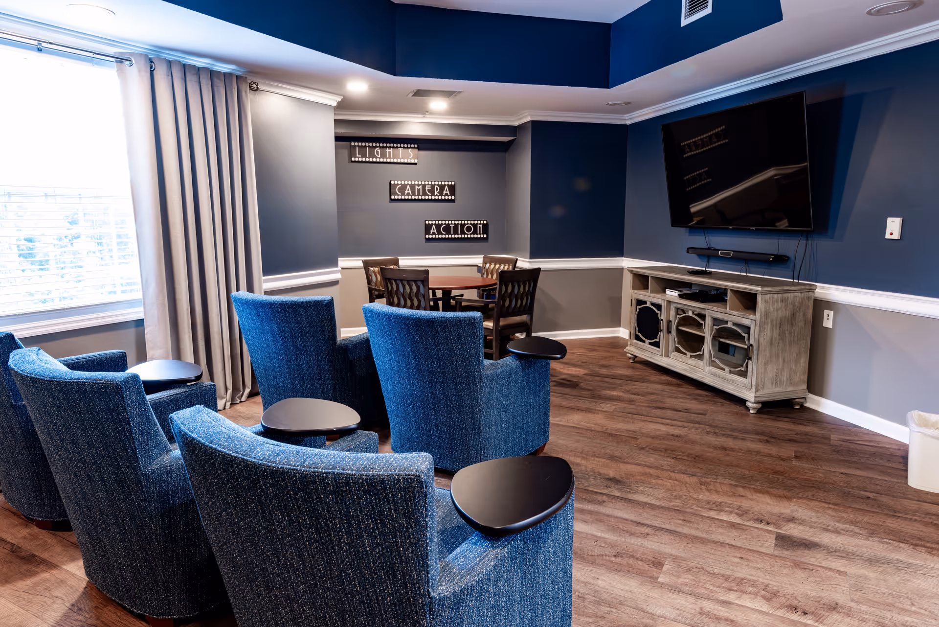 A cozy living room area with four blue upholstered chairs arranged in two rows facing a wall-mounted flat-screen TV. Behind the chairs is a small round table with four wooden chairs. The walls are painted blue and gray with white trim, and there is a large window with beige curtains letting in natural light. Decorative signs on the wall read 'LIGHTS', 'CAMERA', and 'ACTION'.