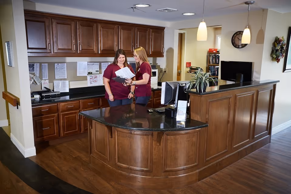 Two women in maroon uniforms standing behind a wooden reception desk with black countertops, reviewing documents together in a well-lit office area with wooden cabinets and a computer monitor.