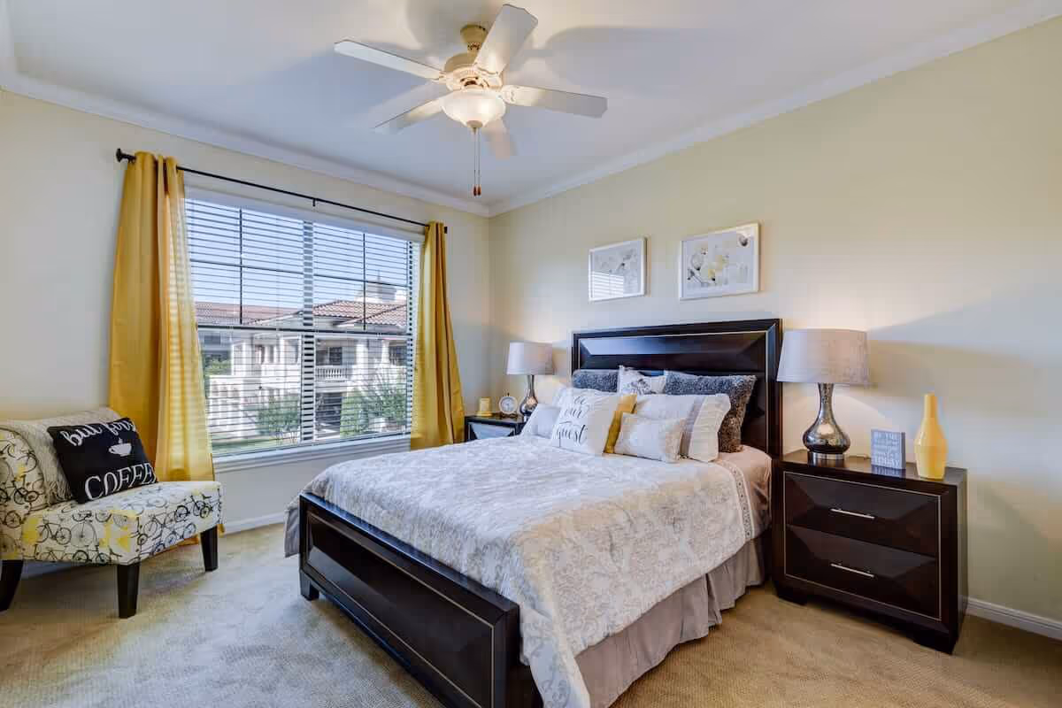 A bright bedroom with a large window covered by yellow curtains, a ceiling fan with light, a dark wooden bed with white and gray bedding and multiple pillows, a matching nightstand with a lamp and decorative items, and a patterned armchair with a pillow that says 'but first COFFEE'.