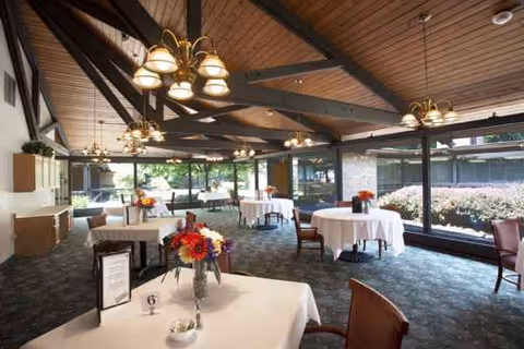 Spacious dining room with round tables, white tablecloths, floral centerpieces, and large windows.