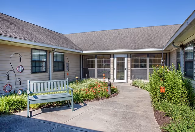 Outdoor courtyard area at a senior living facility with a light blue wooden bench, flower beds with green plants and red flowers, and a concrete pathway leading to a screened porch entrance with a white door and windows.