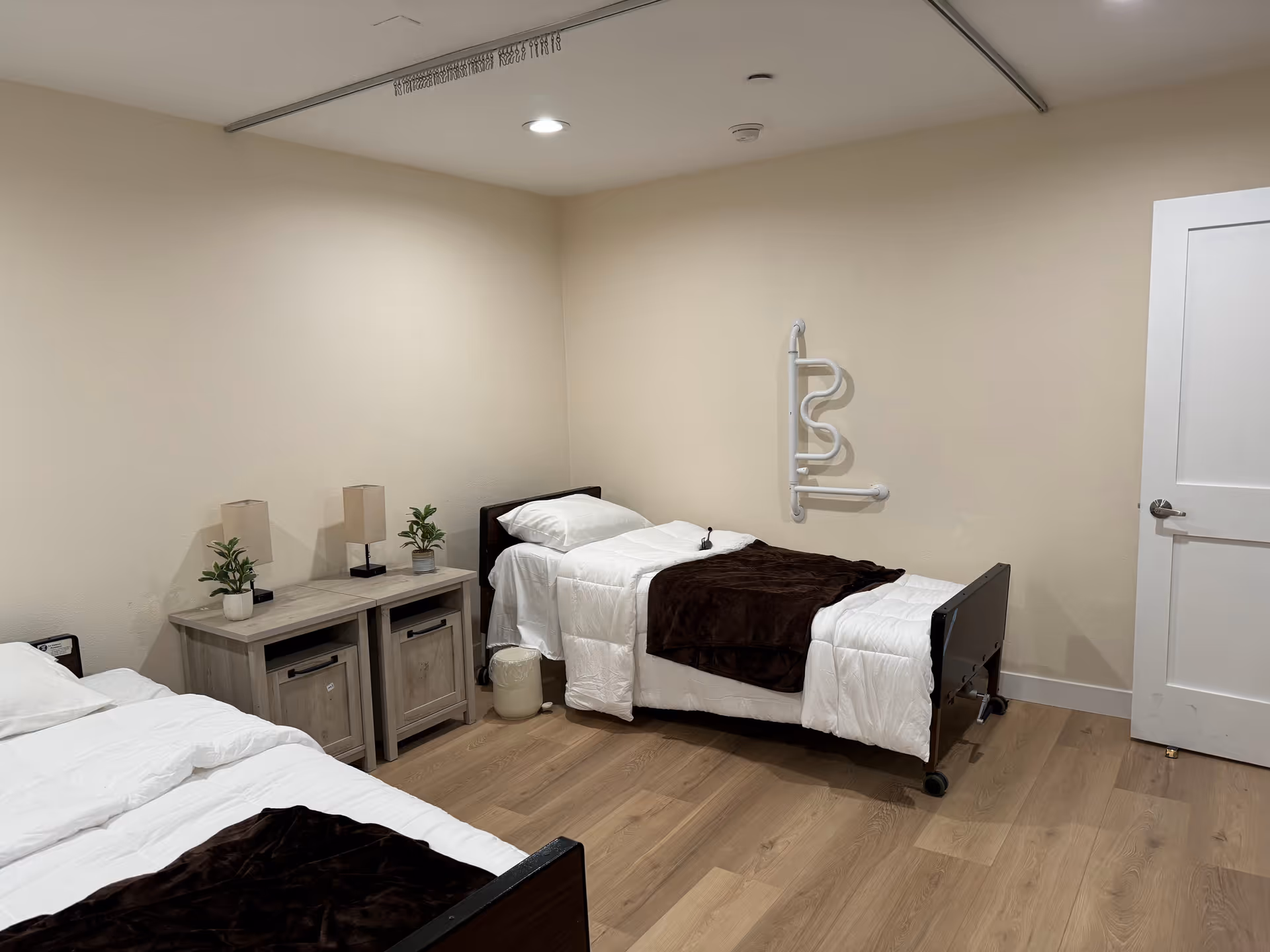 A simple bedroom with two single beds, each with white bedding and a dark brown blanket. Between the beds is a wooden nightstand with two small lamps and two small potted plants. The room has light-colored walls and wood flooring. A white door is visible on the right side of the image, and a white grab bar is mounted on the wall above the bed on the right.
