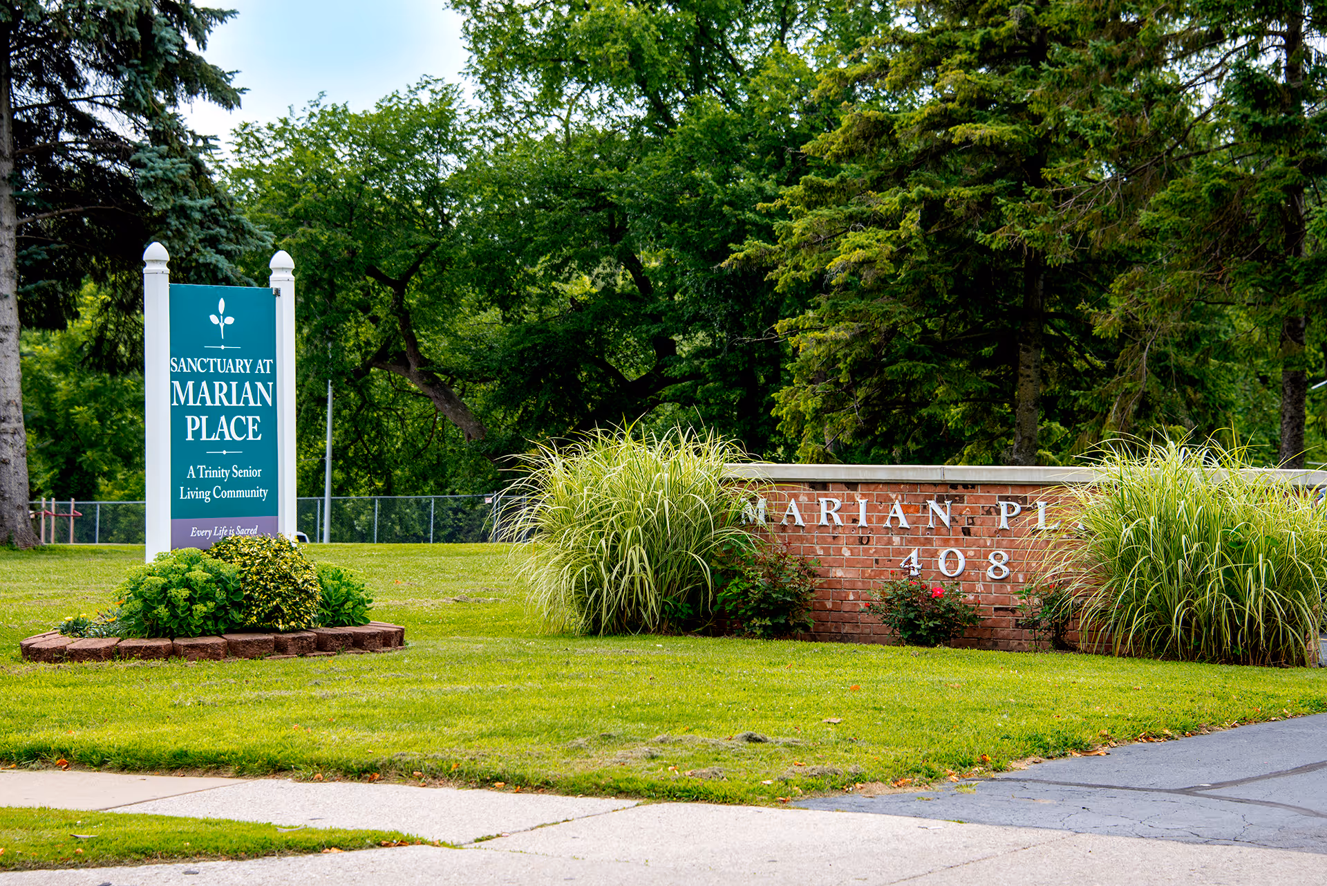 Entrance area of Marian Place senior living community featuring a green sign with white text that reads 'Sanctuary at Marian Place, A Trinity Senior Living Community' and a brick wall with partially visible white letters and the number 408, surrounded by green grass, bushes, and tall trees in the background.