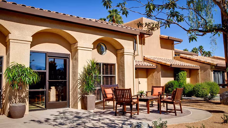 Outdoor patio area at Atria Hacienda with a round concrete seating area featuring four wooden chairs with orange cushions around a small table. The building has beige stucco walls, arched doorways, and tiled roofs. There are potted plants and trees providing shade under a clear blue sky.