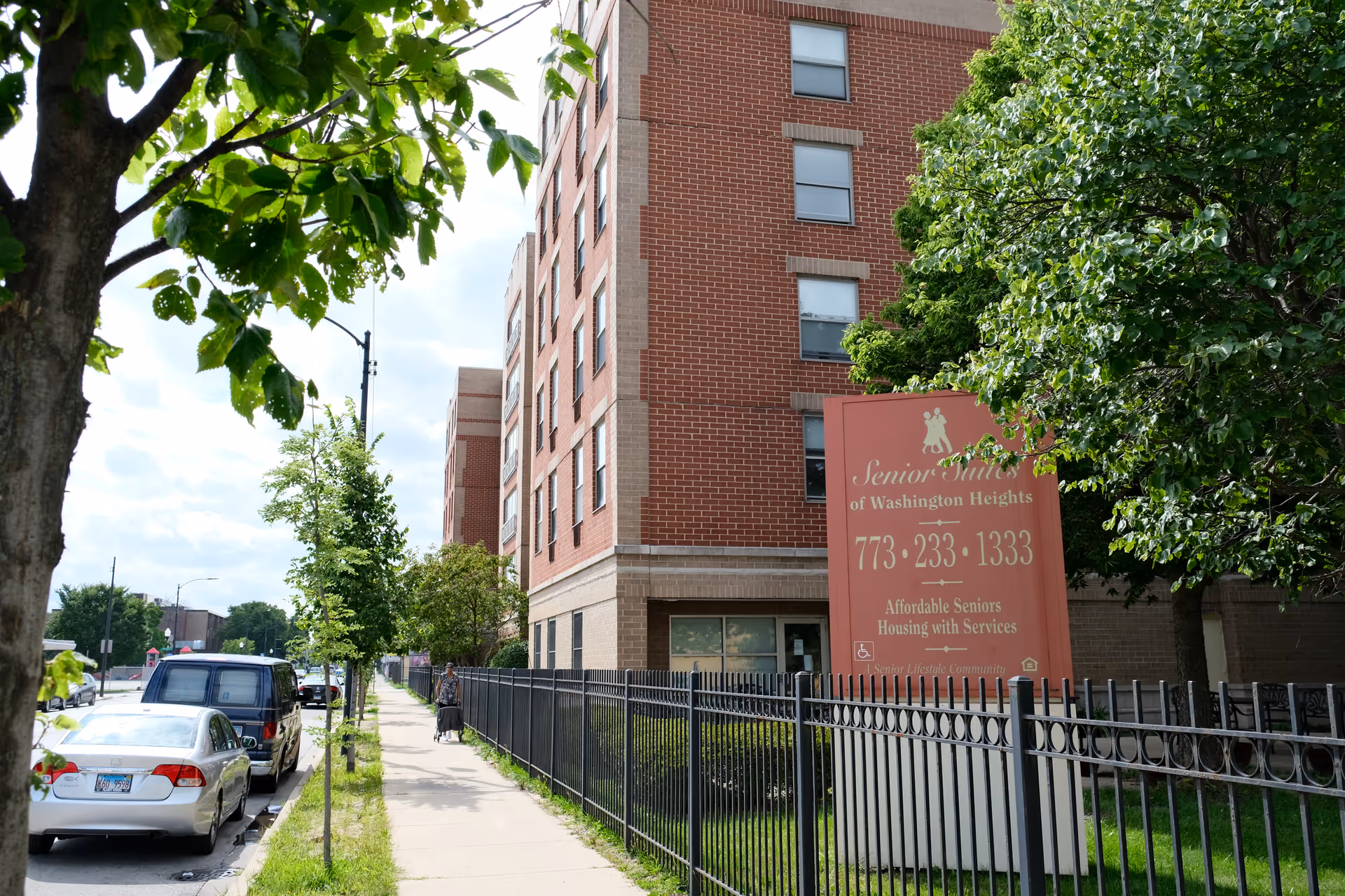 Exterior view of a multi-story brick building with a large red sign in front that reads 'Senior Suites of Washington Heights, 773-233-1333, Affordable Seniors Housing with Services'. There is a black metal fence along the sidewalk, trees with green leaves, parked cars, and a person walking on the sidewalk.