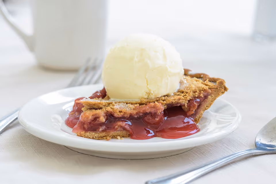 A slice of cherry pie topped with a scoop of vanilla ice cream on a white plate with utensils nearby.