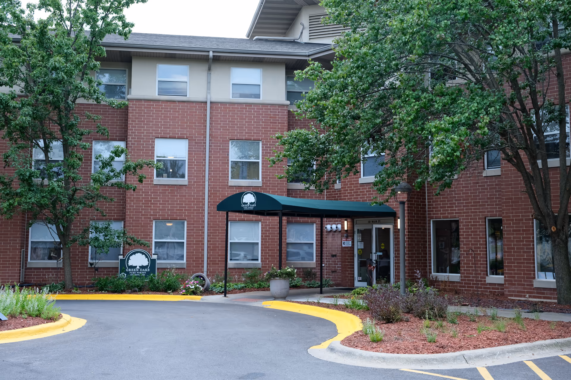 Exterior view of Green Oaks of Park Forest facility showing a red brick building with multiple windows, a green canopy over the entrance, trees, and landscaped areas with plants and mulch.