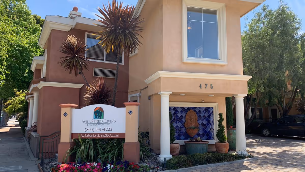 Exterior view of Avila Senior Living at Downtown SLO building with a sign displaying the facility name, phone number, and website. The building is peach-colored with white trim, has two white columns at the entrance, decorative plants, and a tiled water feature. Trees and parked cars are visible in the background.