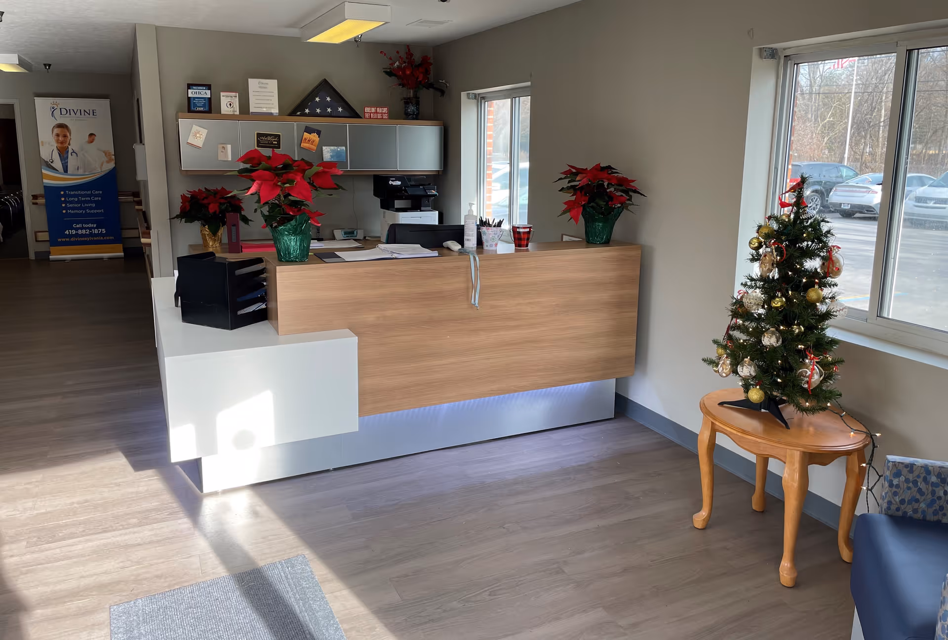 Reception desk and lobby area with poinsettias, a small decorated Christmas tree on a side table, and windows showing parked cars outside.