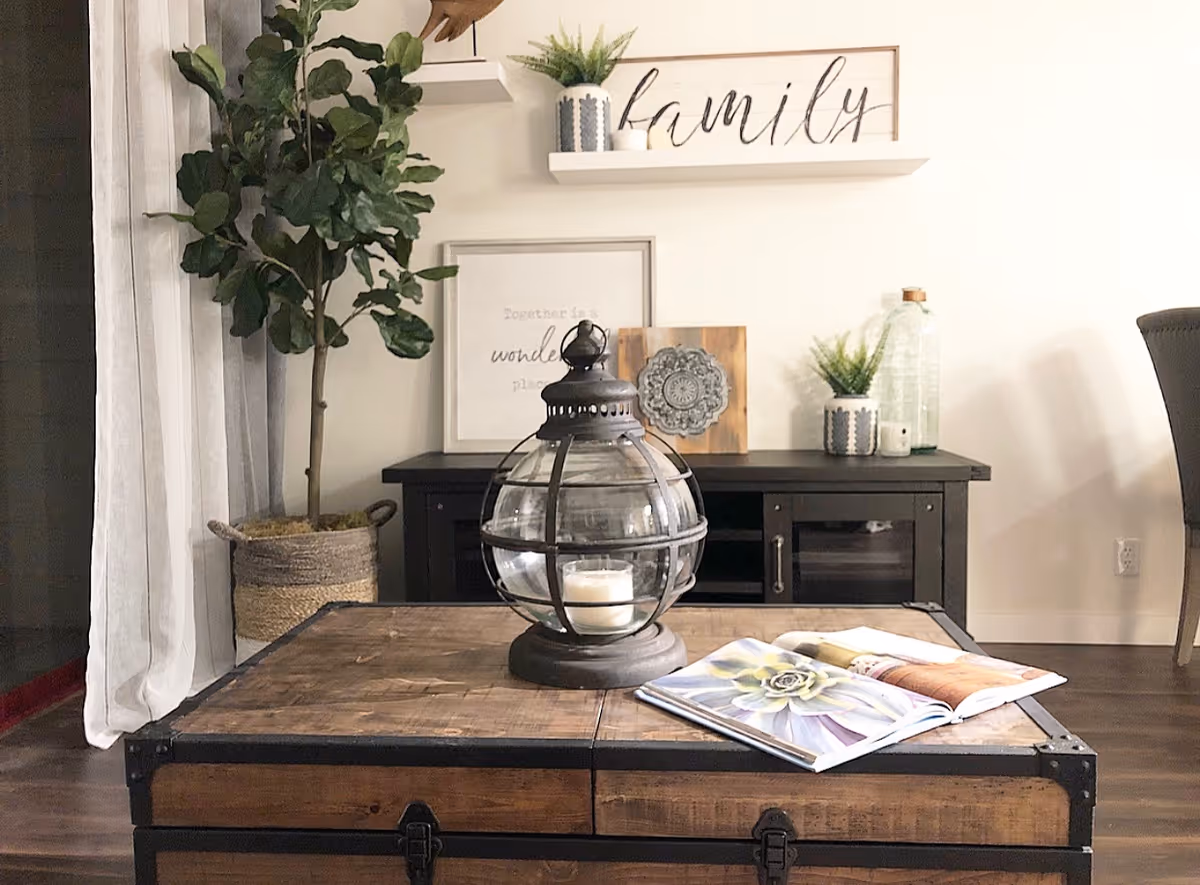 Cozy living room scene with a wooden trunk coffee table topped by a glass lantern and open book, a potted plant, and shelves with decorative items including a 'family' sign.