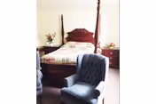 Sunlit bedroom with a four-poster wooden bed flanked by nightstands and two upholstered armchairs in the foreground.