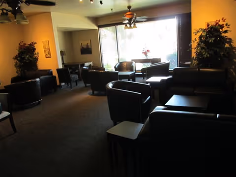 A dimly lit common area with several black leather armchairs and sofas arranged around small tables. Large windows with blinds allow natural light to enter the room. There are ceiling fans with lights, potted plants, and framed artwork on the walls.