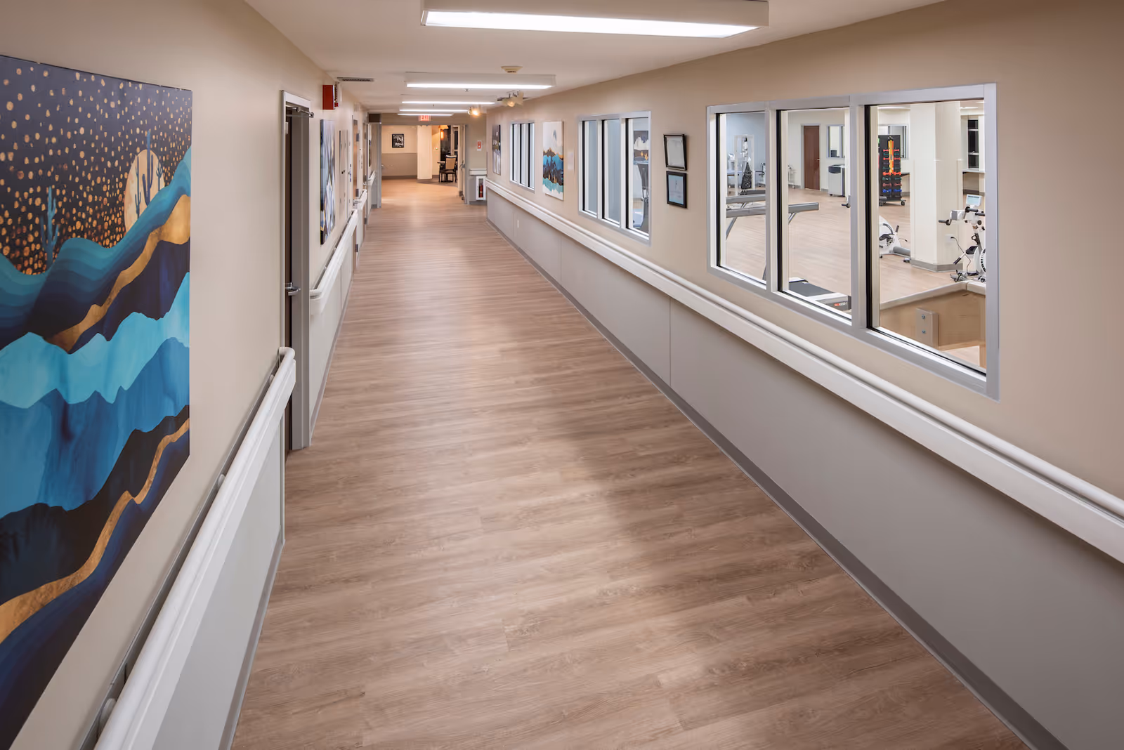 A long, clean hallway in a nursing and rehabilitation center with light wood flooring and beige walls. The left wall features colorful abstract artwork, while the right wall has large windows looking into a fitness or therapy room with exercise equipment. The hallway is well-lit with ceiling lights and has handrails on both sides.