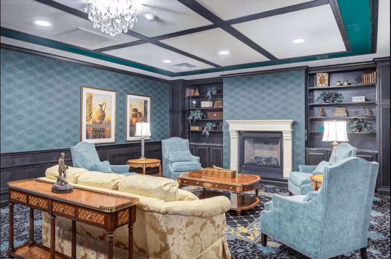A cozy living room with a beige patterned sofa, four blue armchairs, a wooden coffee table, and side tables with lamps. The room features a fireplace with built-in dark wood shelves on either side, decorated with books and plants. Two framed artworks hang on the teal-patterned wall, and a chandelier is visible on the ceiling.