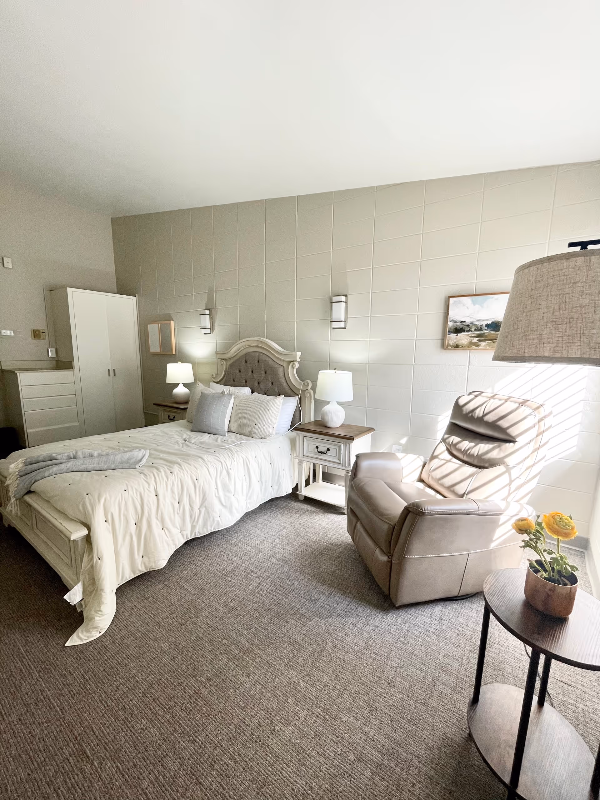 A bright and cozy bedroom with a large bed featuring a tufted headboard, two bedside tables with lamps, a comfortable recliner chair near a window with sunlight streaming in, a small round table with a potted plant, a white wardrobe, and a framed landscape painting on the wall.