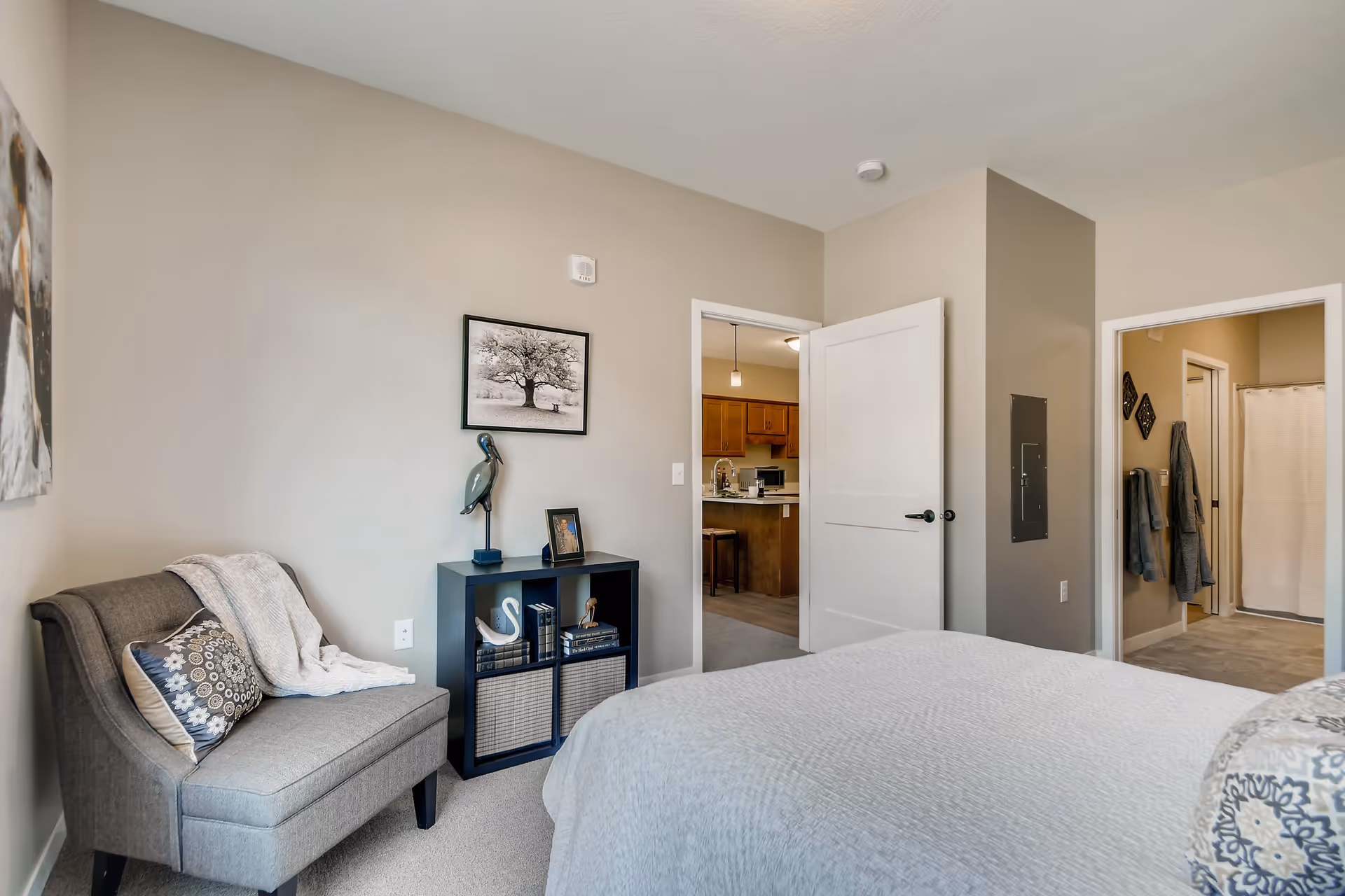A neutral-toned bedroom with a bed, upholstered chair, small shelving unit with decor, and open doors leading to a kitchen and bathroom.