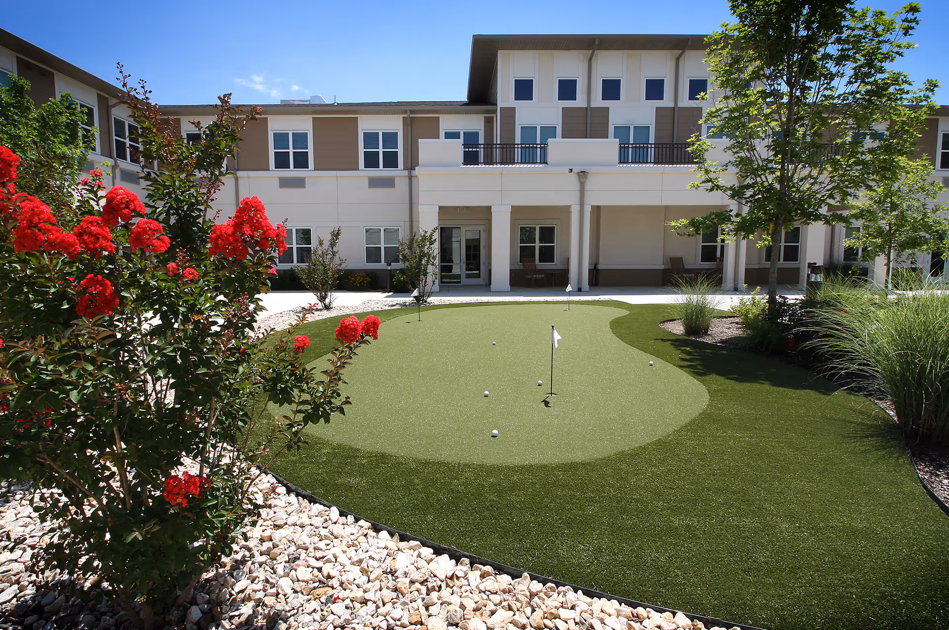 Outdoor putting green with several golf balls and flags surrounded by landscaping with red flowers, bushes, and trees in front of a two-story senior living facility building under a clear blue sky.