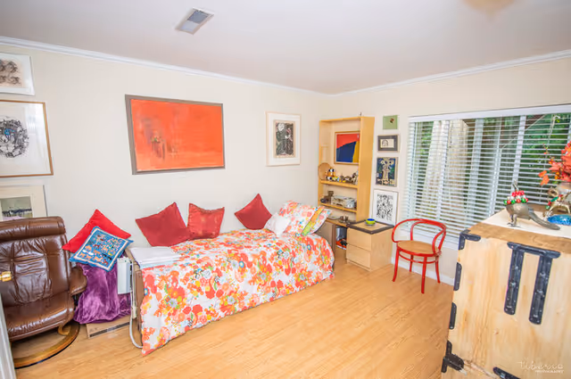 Sunny room featuring a floral-covered daybed with red pillows, a bookshelf, leather chair and a window with blinds.