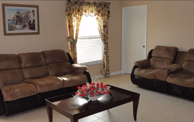 A living room with two brown cushioned sofas facing each other, a wooden coffee table with a flower arrangement in the center, a window with floral curtains, a closed white door, and a framed picture on the wall.