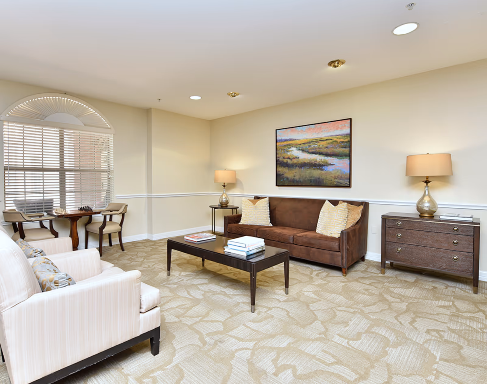 A cozy living room with a brown sofa adorned with two patterned pillows, a dark wooden coffee table with books on it, two beige armchairs with patterned cushions, a small round table with two chairs near a window with blinds, two table lamps on side tables, and a landscape painting on the wall.