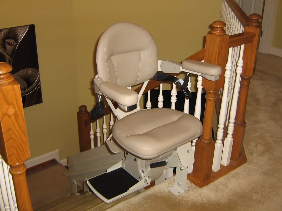 Beige stairlift chair installed at the top of a carpeted staircase next to a wooden banister.