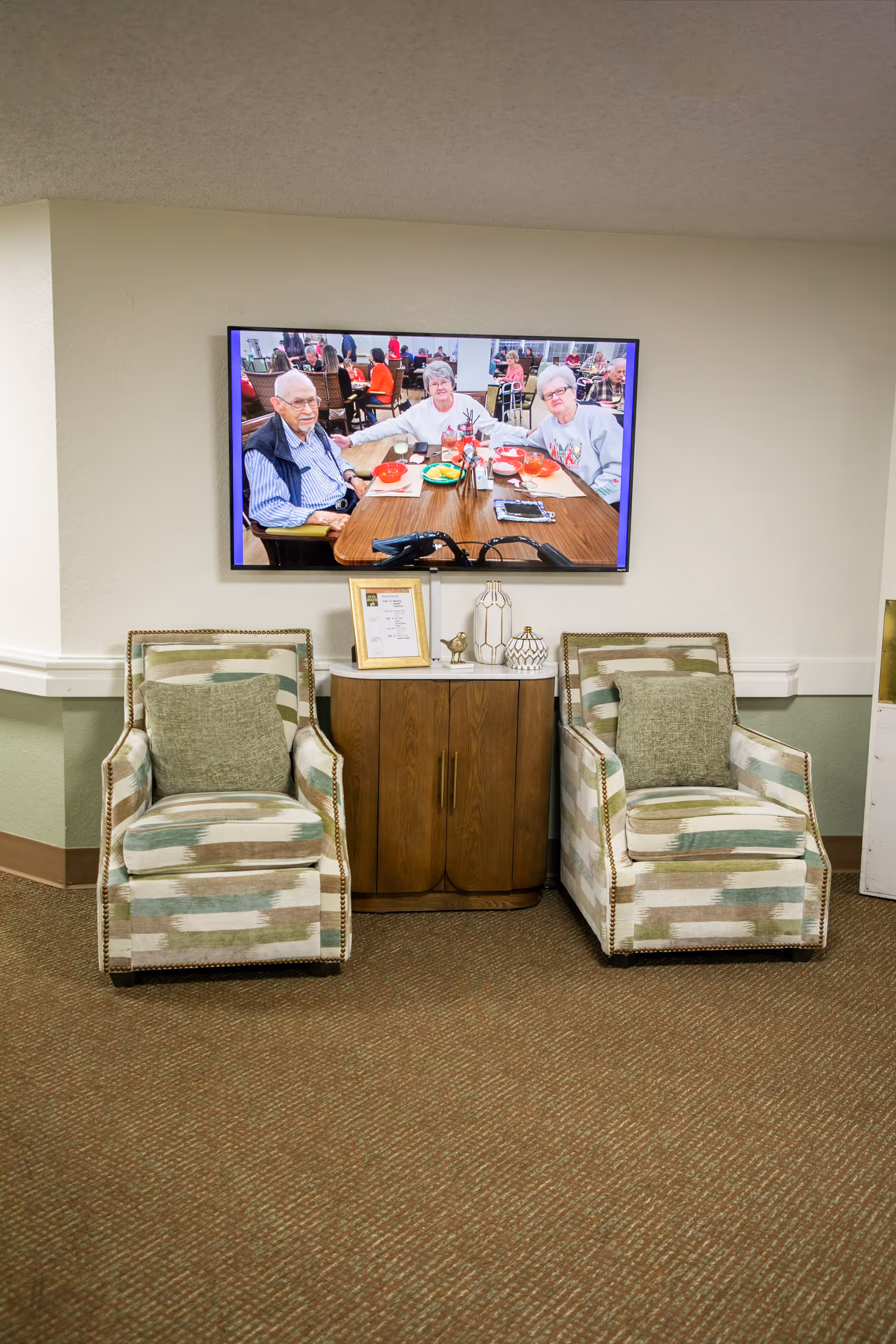 Two patterned armchairs with green cushions are placed on either side of a wooden cabinet with decorative items on top. Above the cabinet is a wall-mounted TV displaying an image of elderly people sitting at a dining table in a communal setting.
