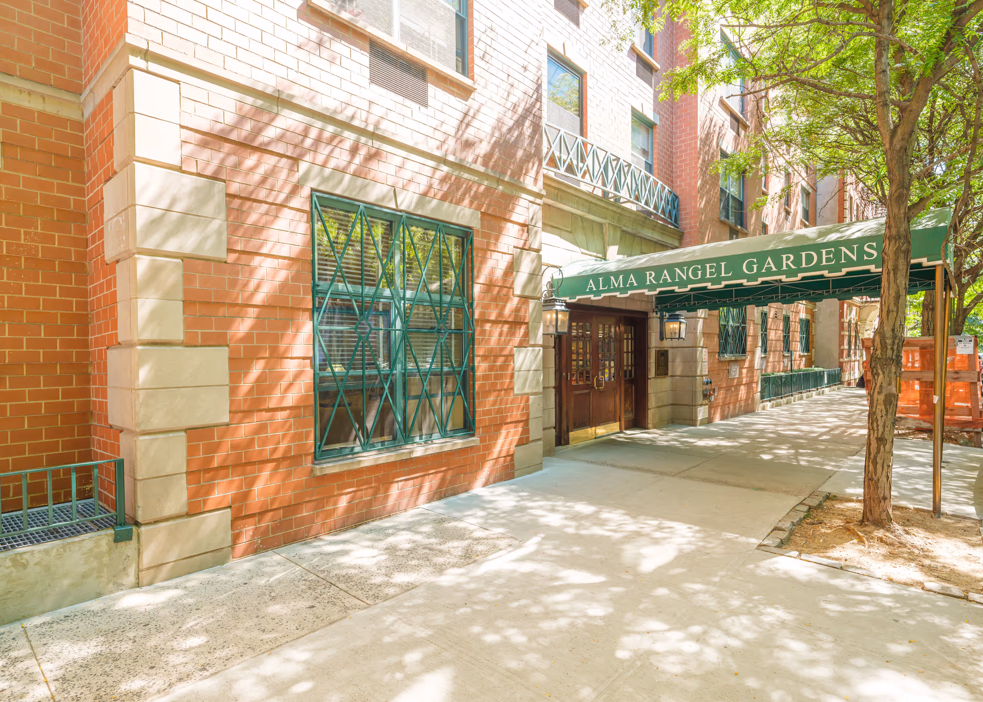 Exterior view of a brick building with green window grilles and a green awning above the entrance that reads 'ALMA RANGEL GARDENS'. There are trees casting shadows on the sidewalk in front of the building.