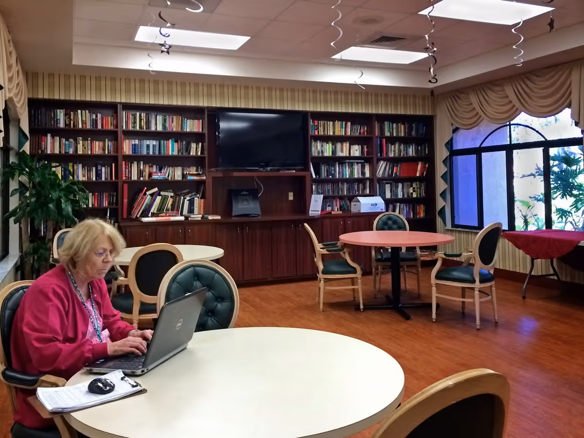 An elderly woman works on a laptop at a round table in a library-style common room with bookshelves and a TV.