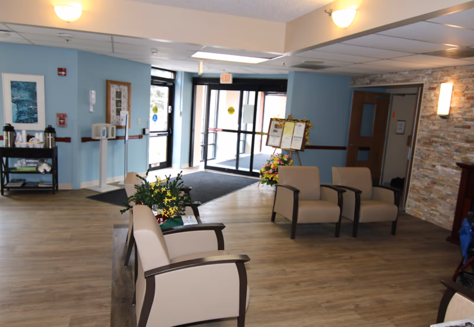 A senior living lobby with beige armchairs, potted plants, and glass double entrance doors.