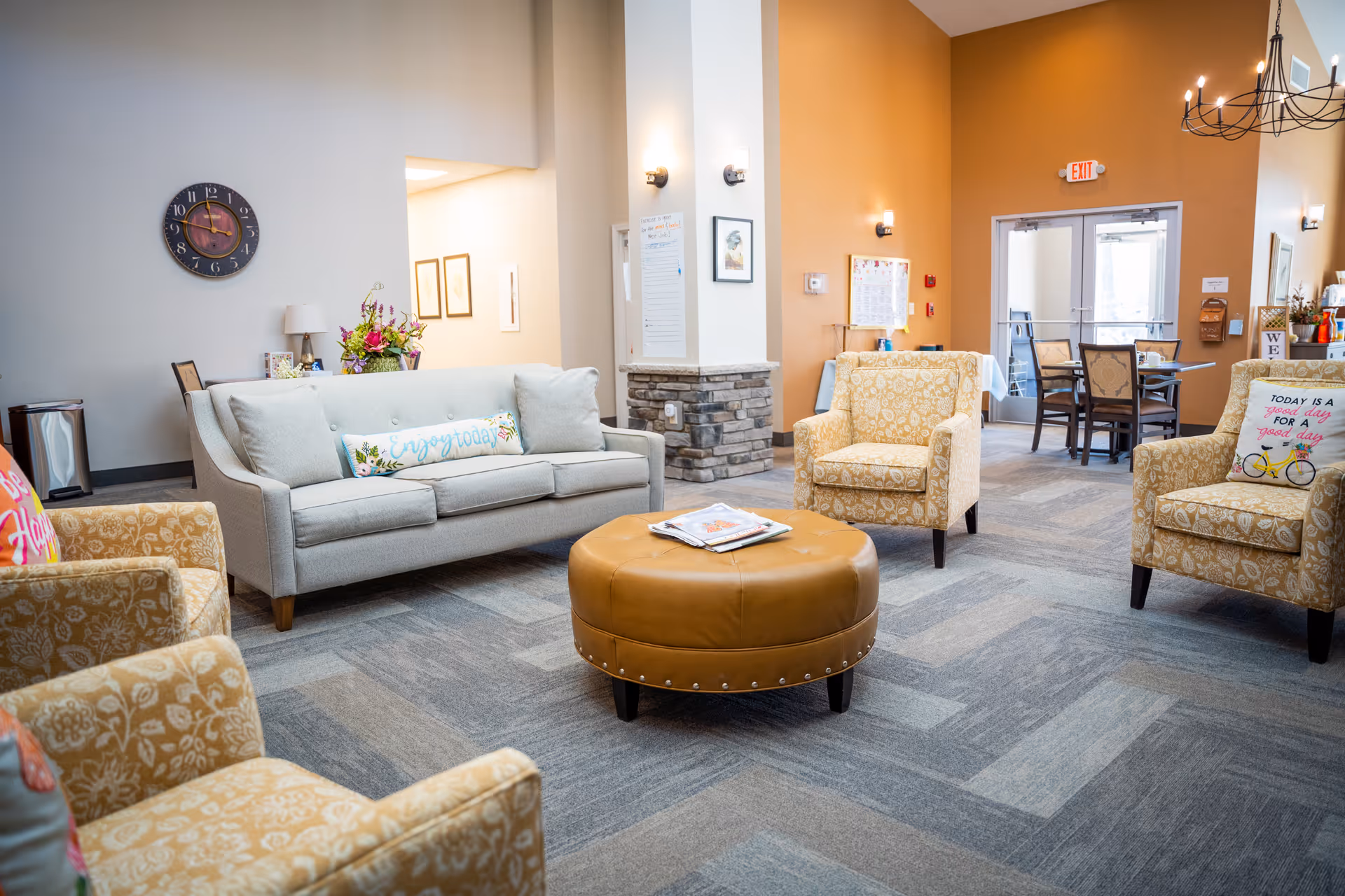 A bright and welcoming assisted living facility common area with a light gray sofa adorned with a decorative pillow that says 'Enjoy today', surrounded by four patterned armchairs. In the center is a round, tan leather ottoman with magazines on top. The room features a mix of light gray and warm orange walls, a wall clock, framed artwork, and a chandelier. In the background, there is a dining area with tables and chairs near glass doors.