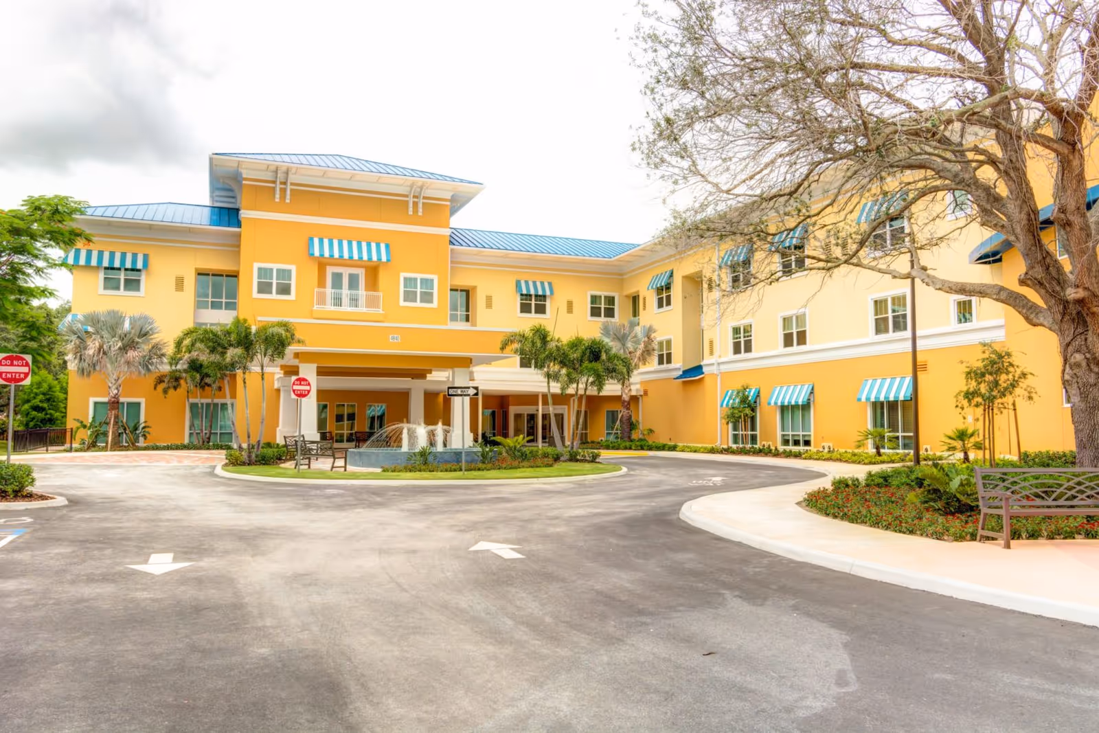 Yellow three-story building with teal-striped awnings, a circular driveway, palm trees, benches and a fountain at the main entrance.