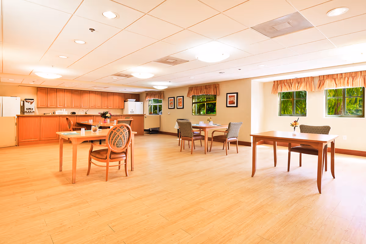 A bright and spacious dining area with wooden floors and several tables and chairs arranged for seating. The room features large windows with curtains, allowing natural light to fill the space. The back wall has a long counter with wooden cabinets above and below, along with a refrigerator and coffee machine. The walls are decorated with framed artwork.