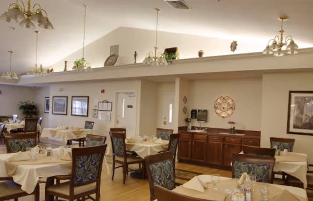 A dining room in a senior living facility with several tables covered in beige tablecloths, each set with napkins, glasses, and silverware. The room has wooden chairs with patterned upholstery, light-colored walls, framed artwork, and multiple chandeliers hanging from the ceiling. There is a wooden sideboard against one wall with decorative items and a water dispenser.