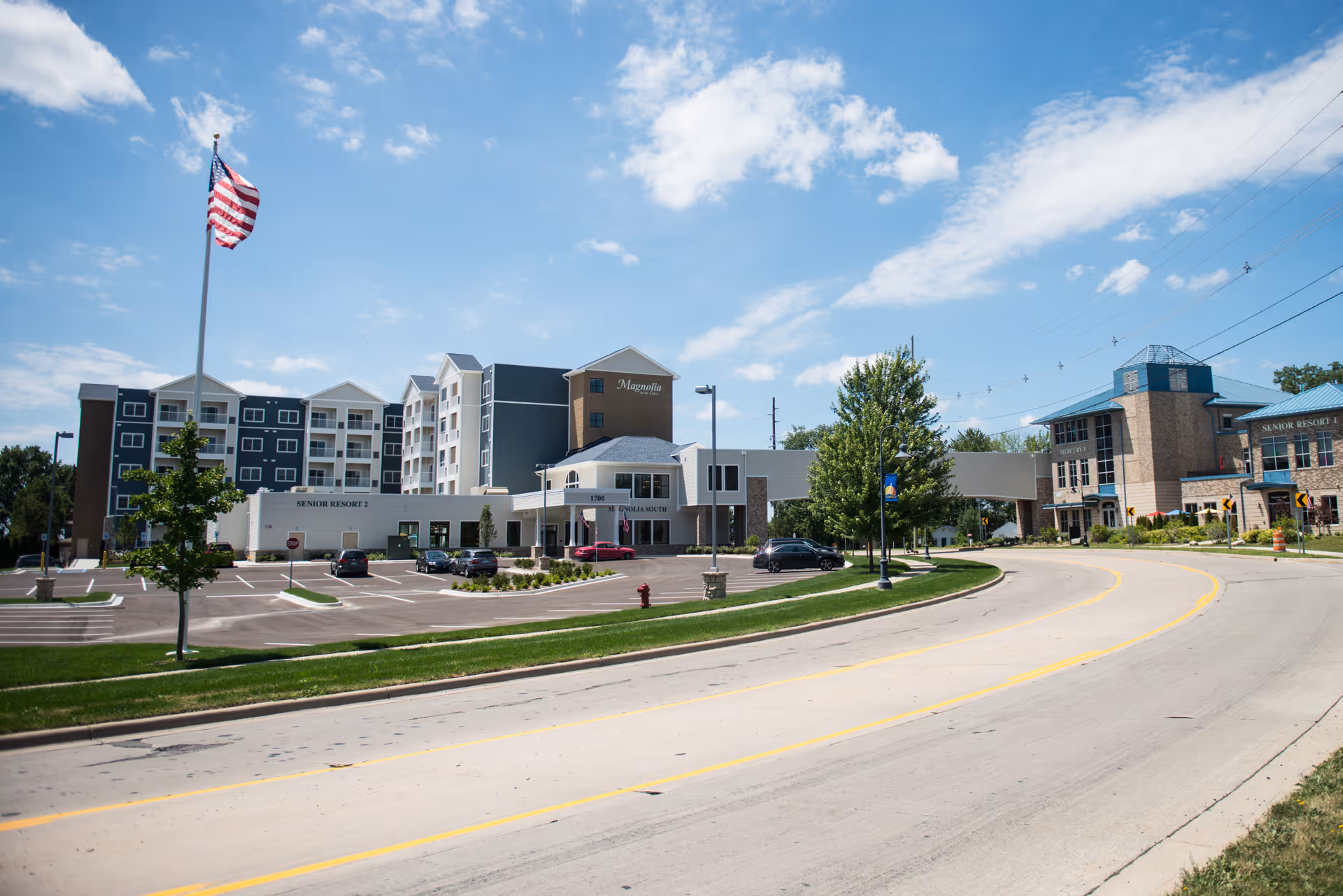 Exterior view of Magnolia by the Lakes senior living facility showing multiple connected buildings with a parking lot, an American flag on a tall flagpole, trees, and a curved road in the foreground under a partly cloudy blue sky.